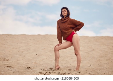 Beautiful Woman With Windy Hair Standing On Sandy Beach. Stylish Young Sexy Female In Knitted Sweater And Bare Legs Relaxing On Coast.