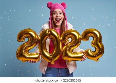 Beautiful woman in winter coat holding gold colored numbers and smiling against blue background - Shutterstock ID 2220201685