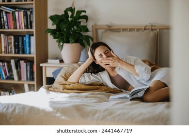 Beautiful woman yawning, reading book in bed, cozy, warm and contented feeling. Freedom in story, weekend activity for single woman. - Powered by Shutterstock