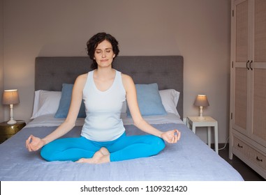 Beautiful Woman In Yoga Position On Bed In Home Bedroom Interior At Night, Meditating With Eyes Shut. Spiritual Activities Early Morning, Indoors. Leisure Recreation Activities, Serene Lifestyle.