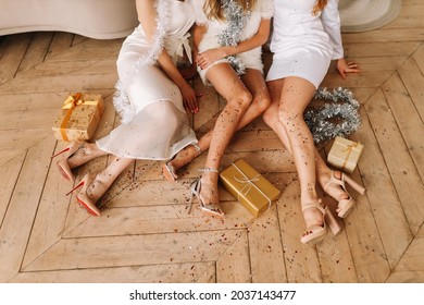 Beautiful women in elegant dresses with long legs in fashionable heels sit on the wooden floor among confetti and gift boxes celebrate Christmas and new year at a house party, a selective focus