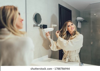 Beautiful women enjoying in hotel room.Young happy smiling woman wearing bathrobe using hair dryer in bathroom.