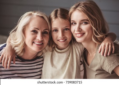 Beautiful women generation: granny, mom and daughter are hugging, looking at camera and smiling while sitting on couch at home