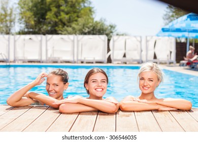 Beautiful Women Are Standing In Swimming Pool And Resting. They Are Leaning On The Board With Their Hands. The Ladies Are Looking At The Camera And Laughing