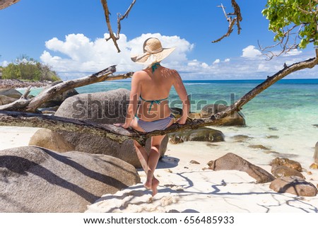 Beautiful women wearing bikini and sun hat sitting on tree branch at picture perfect tropical beach on Mahe island, Seychelles, looking at emerald sea and white clouds on horizon.