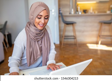 Beautiful Women Wearing The Islamic Headscarf, Sitting On A Wooden Table Holding Pencil Writing On Notebook And Cute Smile With Computer. Attractive Female Arabic Working On Laptop Computer 