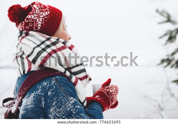 Beautiful women wearing in red hat, mittens and a\
cozy scarf stands near the winter lake and holds a cup of warm tea\
in her hands