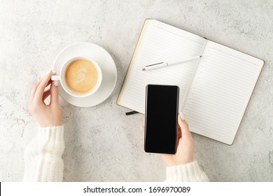 Beautiful women's hands holding up-to-date smartphone and writing in notepad with cup of coffee on concrete background. Women's business concept. Coffee break top view. Mockup for your design. - Powered by Shutterstock