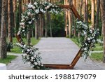 A beautiful wooden wedding arch in the form of a hexagon, decorated with natural flowers, stands on the road against the background of a forest with pine trees.