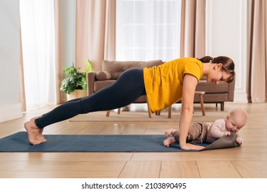 Beautiful yogi mom smiling at her baby while doing push up on exercising mat at home. Happy mom working out with her son. Young mom looking at her little baby during her post-natal fitness routine