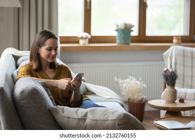Beautiful young 30s woman holds smartphone smile read pleasant message, enjoy new mobile application relaxing alone on sofa. Buying goods e-services on internet, making order, do e-shopping at home Foto Stock