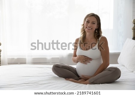 Beautiful young 35s pregnant woman in comfort home-wear touch big belly in late pregnancy smile look at camera sit on bed in light bedroom at home. Happy expectant mother portrait, maternity concept