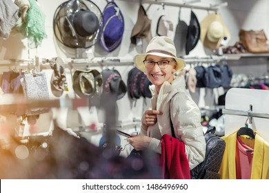 Beautiful Young Adult Caucasian Bald Woman Choosing And Trying On Hat In Department Retail Store At Shopping Mall. Portrait Of Happy Girl During Shopping