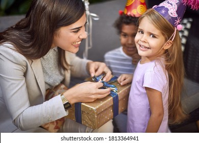 Beautiful Young Adult Female Unwrapping Birthday Present For Little Caucasian Girl Looking At Camera, Smiling