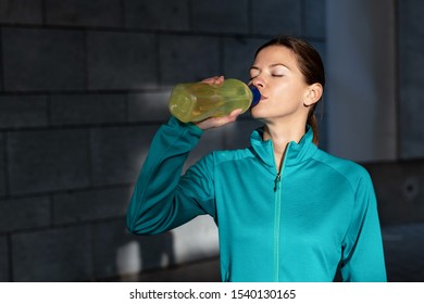 Beautiful young adult sportswoman in sportswear drinking refreshing water from plastic bottle and resting outdoor after running training in city. Athletic girl standing near wall with copy space - Shutterstock ID 1540130165