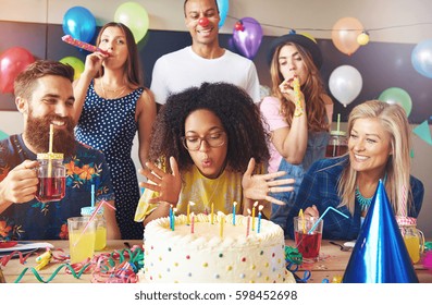 Beautiful Young Adult Woman Blowing Out Candles On Her Cake At Birthday Party Surrounded By Celebrating Friends