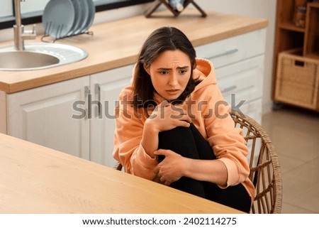 Beautiful young afraid woman sitting at table in kitchen