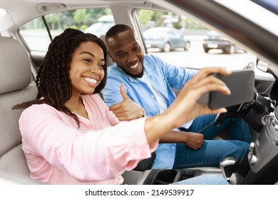Beautiful young African American couple taking selfie inside car, using mobile phone, showing thumbs up sign gesture. Happy black man and woman going on vacation, taking self photo, buying new auto