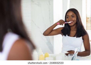 Beautiful Young African American Female Brushing Her Teeth With Toothbrush In Bathroom, Happy Black Woman Smiling At Mirror, Enjoying Making Morning Hygiene At Home, Selective Focus On Reflection