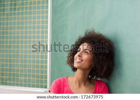 Beautiful young African American student with a wild afro hairstyle standing daydreaming in front of the blackboard smiling as she allows her imagination free reign
