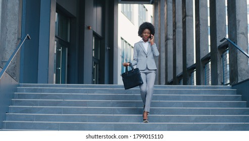 Beautiful Young African American Woman Walking And Talking On Mobile Phone At Steps Down From Business Center. Female Speaking On Cellphone And Stepping Down. Businesswoman Chatting In Telephone Call.