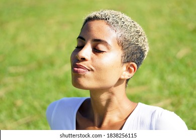 Beautiful Young African American Woman Sits On Grass At The Park And Soaks Up The Sun's Rays With Eyes Closed And Short Hair Outside                              