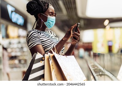 Beautiful young African American woman wearing a medical protective mask on her face holding shopping bags using a mobile phone in a mall while shopping in a women's boutique, COVID-19 quarantine