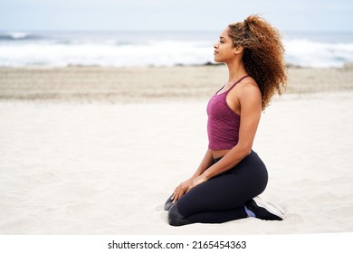 Beautiful Young African American Woman Wearing Workout Clothing Of Leggings And Crop Top Kneels On The Sand At The Beach For A Side View Portrait                              