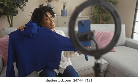 A beautiful young african american woman with curly hair holds up a blue sweater while recording a video in her living room, featuring cozy home decor and indoor plants. - Powered by Shutterstock