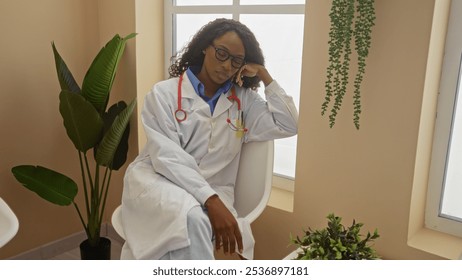 A beautiful young african american woman with curly hair, dressed in a white doctor's coat, is sitting in a hospital waiting room environment, appearing thoughtful and relaxed. - Powered by Shutterstock