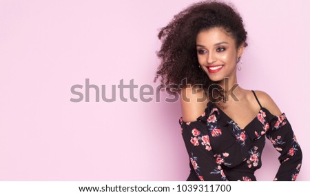 Beautiful young african american woman with afro hairstyle posing on pink pastel background wearing fashionable floral dress. Girl smiling, looking at camera.