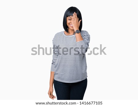 Beautiful young african american woman wearing stripes sweater over isolated background covering one eye with hand with confident smile on face and surprise emotion.