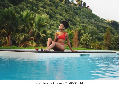 A Beautiful Young African Descent Woman Relaxing Poolside Drinking Healthy Fruit Juice - African American Girl Sunbathing By The Swimming Pool - Travel And Vacation Concept