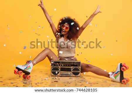 Beautiful young african woman with afro hairstyle throwing confetti, showing peace gesture while sitting in roller skates with boombox, isolated on yellow background