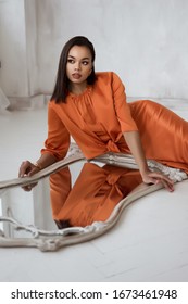 Beautiful Young African Woman Lying Under Mirror. Mirror Reflection. Fashion Model Posing On The Floor. Trendy Orange Dress. Stylish Outfit.  Indoor, Studio Shot. Woman In Elegant Evening Dress 