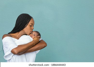 A beautiful young African-American woman with braids is kissing her newborn son and looking at him with love on a teal blue background.
