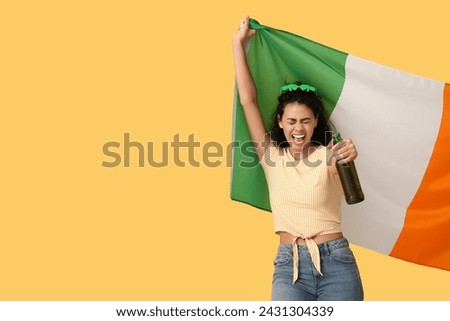 Beautiful young African-American woman with Irish flag and bottle of beer on yellow background. St. Patrick's Day celebration