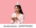 Beautiful young African-American woman in party hat holding birthday cupcake with burning candle on pink background