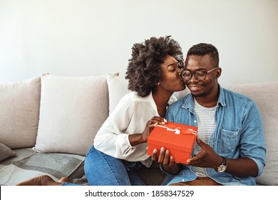 Beautiful Young Afro American Couple On Couch At Home. Handsome Guy Giving A Gift Box. Happy African American Couple Celebrating Winter Holidays Together