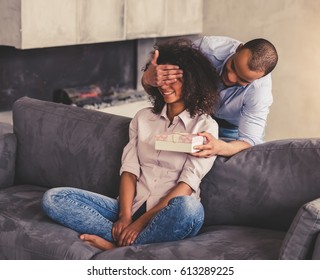 Beautiful Young Afro American Couple On Couch At Home. Handsome Guy Is Covering His Girlfriend Eyes And Giving A Gift Box