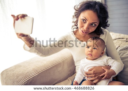 Beautiful young Afro American mother is making selfie with her adorable little baby and smiling while sitting on sofa at home