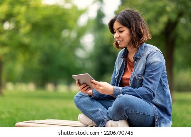 Beautiful Young Arab Female Student Using Digital Tablet While Sitting On Bench Outdoors, Smiling Middle Eastern Woman Relaxing Outside With Modern Gadget, Browsing Internet On Tab Computer