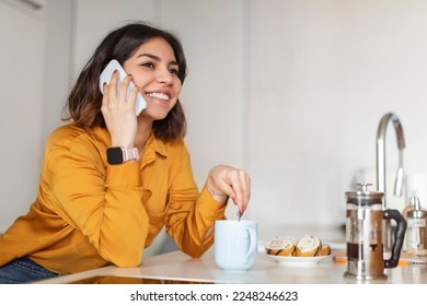 Beautiful Young Arab Female Talking On Cellphone And Drinking Coffee In Kitchen At Home, Smiling Middle Eastern Woman Enjoying Mobile Call, Having Pleasant Cellphone Conversation With Friend - Powered by Shutterstock