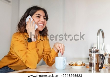 Beautiful Young Arab Female Talking On Cellphone And Drinking Coffee In Kitchen At Home, Smiling Middle Eastern Woman Enjoying Mobile Call, Having Pleasant Cellphone Conversation With Friend