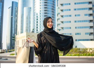 Beautiful Young Arab Muslim Woman In Abaya Clothes Holding Shopping Bags And Walking On The City Street. Shopping Time. Modern Skyscrapers In The Background.