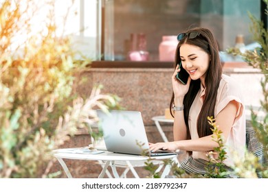 Beautiful Young Asian Business Woman Talking Cellphone While Typing At The Laptop. Remote Work Outside The Office, Outdoor Workplace.