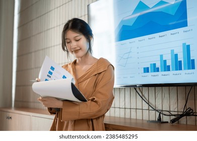 A beautiful young Asian businesswoman or female financial analyst is reading information in her paperwork while presenting her financial analysis information in the meeting. - Powered by Shutterstock