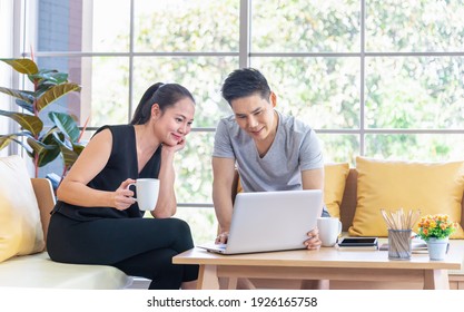 Beautiful young asian couple using laptop on the sofa at home. Young man working from home on laptop. Woman is smiling at her. Happy couple enjoying work from home
