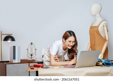 Beautiful young Asian fashion designer or tailor working with laptop computer to search idea, inspiration or online orders, full of tailoring tools with mannequin and equipment on desk in studio