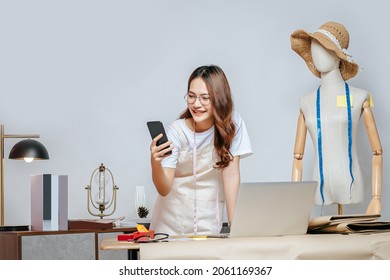 Beautiful young Asian fashion designer or tailor with working with laptop computer and use smartphone during works, full of tailoring tools  with mannequin and equipment on the desk in the studio